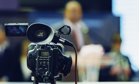 Television camera recording publicity event. Camera in focus, spokesman blurred